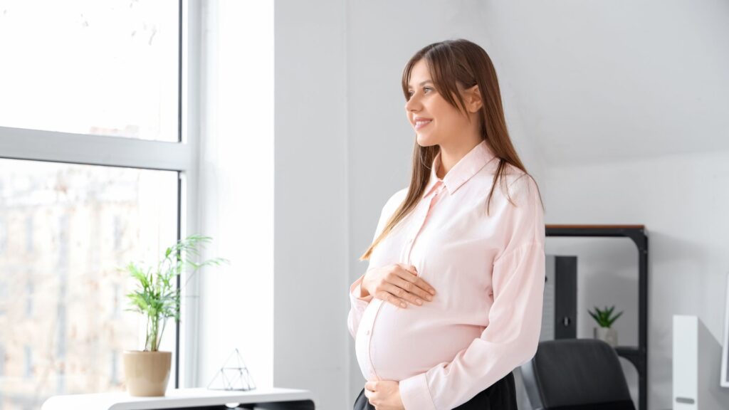 A brown-haired person in smart clothing holds their pregnancy bump and smiles.